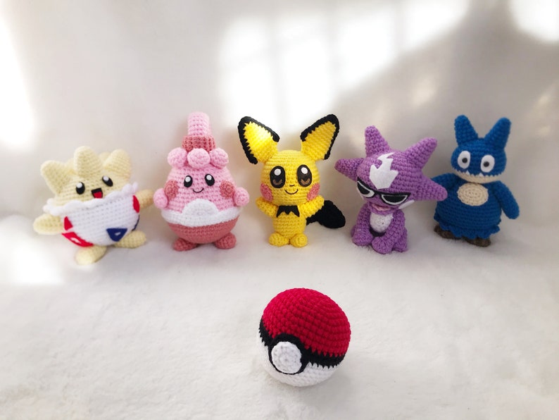 May include: A collection of six handmade crochet Pok&eacute;mon figures, including Pikachu, in various colors like yellow, pink, purple, and blue. A red and white Pok&eacute; Ball is in the foreground. The figures are arranged on a white surface.