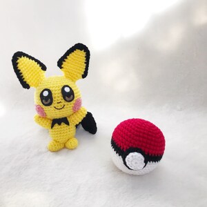 May include: A handmade yellow Pikachu amigurumi with black-tipped ears, a black tail, and pink cheeks, next to a red and white crocheted Pok&eacute; Ball. The plush toys are on a white surface.