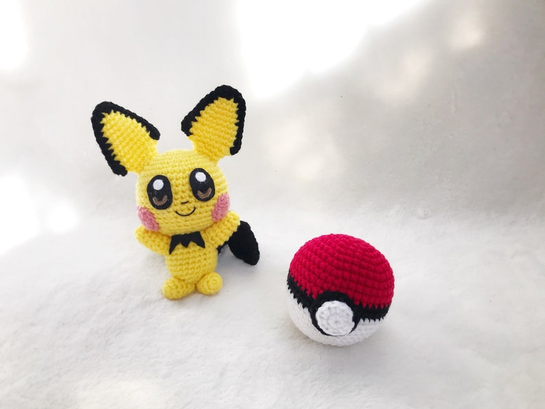 May include: A handmade yellow Pikachu amigurumi with black-tipped ears, a black tail, and pink cheeks, next to a red and white crocheted Pok&eacute; Ball. The plush toys are on a white surface.