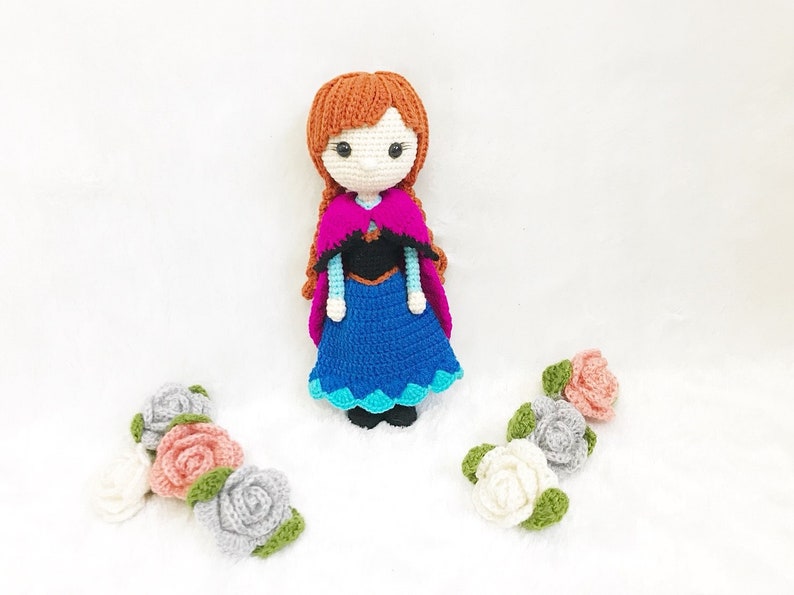 May include: A crocheted doll dressed as Anna from the movie Frozen. The doll has brown hair, a blue dress, a pink cape, and a black bodice. The doll is surrounded by crocheted flowers in shades of pink, white, and gray.
