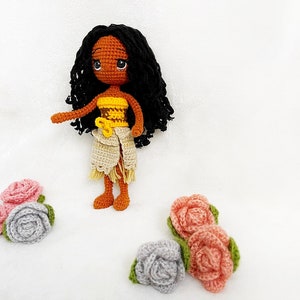 May include: A crocheted doll with brown skin, black curly hair, and a yellow and brown top. The doll is wearing a white skirt and is standing on a white surface. There are several crocheted flowers in pink and gray on the surface.