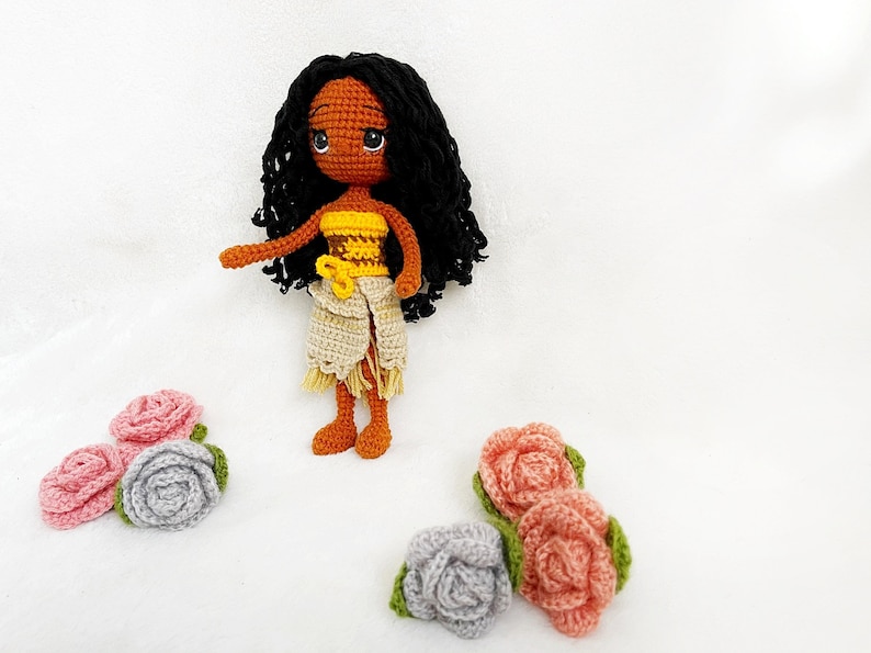 May include: A crocheted doll with brown skin, black curly hair, and a yellow and brown top. The doll is wearing a white skirt and is standing on a white surface. There are several crocheted flowers in pink and gray on the surface.