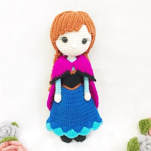 May include: Crocheted doll with brown hair, a blue and teal dress, a black bodice, and a pink and black cape.
