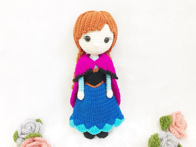 May include: Crocheted doll with brown hair, a blue and teal dress, a black bodice, and a pink and black cape.