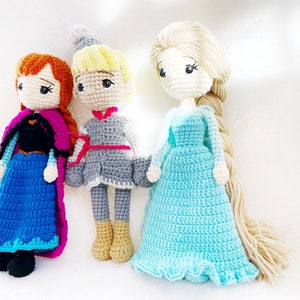 May include: Three crocheted dolls, one with a blue dress and a pink cape, one with a grey dress and a red sash, and one with a light blue dress and long blonde hair.