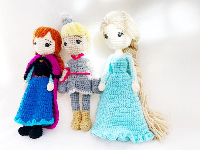 May include: Three crocheted dolls, one with a blue dress and a pink cape, one with a grey dress and a red sash, and one with a light blue dress and long blonde hair.