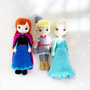 May include: Three crocheted dolls, a girl with red hair wearing a blue and purple dress, a boy with blonde hair wearing a grey and red outfit, and a girl with blonde hair wearing a light blue dress.