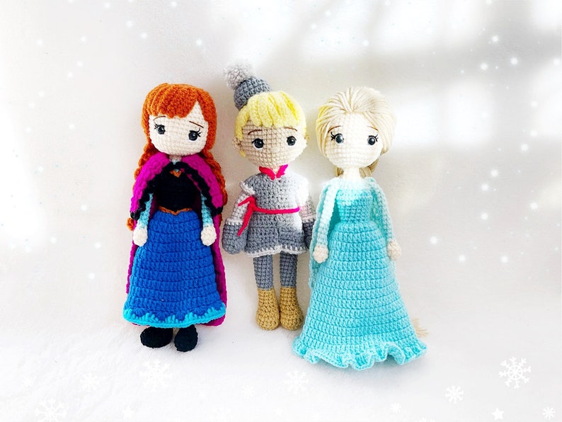 May include: Three crocheted dolls, a girl with red hair wearing a blue and purple dress, a boy with blonde hair wearing a grey and red outfit, and a girl with blonde hair wearing a light blue dress.