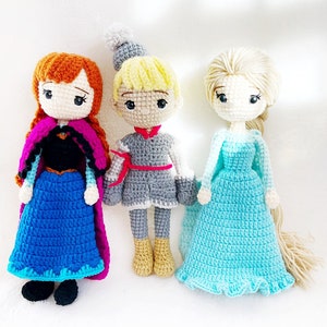 May include: Three crocheted dolls, one with a blue dress and a pink cape, one with a grey dress and a red sash, and one with a light blue dress and long blonde hair.