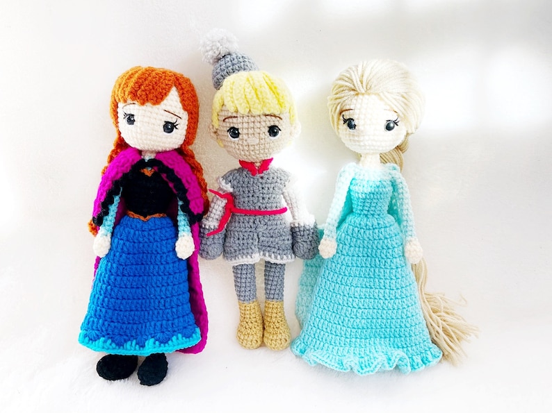 May include: Three crocheted dolls, one with a blue dress and a pink cape, one with a grey dress and a red sash, and one with a light blue dress and long blonde hair.