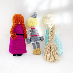 May include: Three crocheted dolls standing back to back. The doll on the left has long, braided, orange hair and is wearing a purple dress. The doll in the middle has short, blonde hair and is wearing a grey dress with a red belt. The doll on the right has long, braided, blonde hair and is wearing a light blue dress.