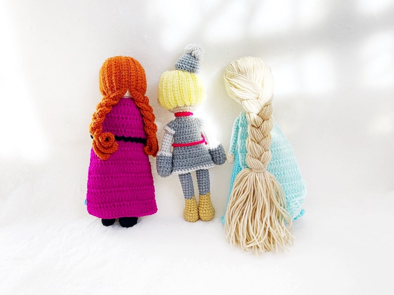May include: Three crocheted dolls standing back to back. The doll on the left has long, braided, orange hair and is wearing a purple dress. The doll in the middle has short, blonde hair and is wearing a grey dress with a red belt. The doll on the right has long, braided, blonde hair and is wearing a light blue dress.