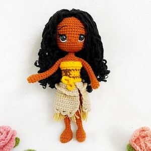 May include: Crocheted doll with brown skin, black curly hair, and a yellow and brown top. The doll is wearing a beige skirt with a fringe and is standing on a white background.