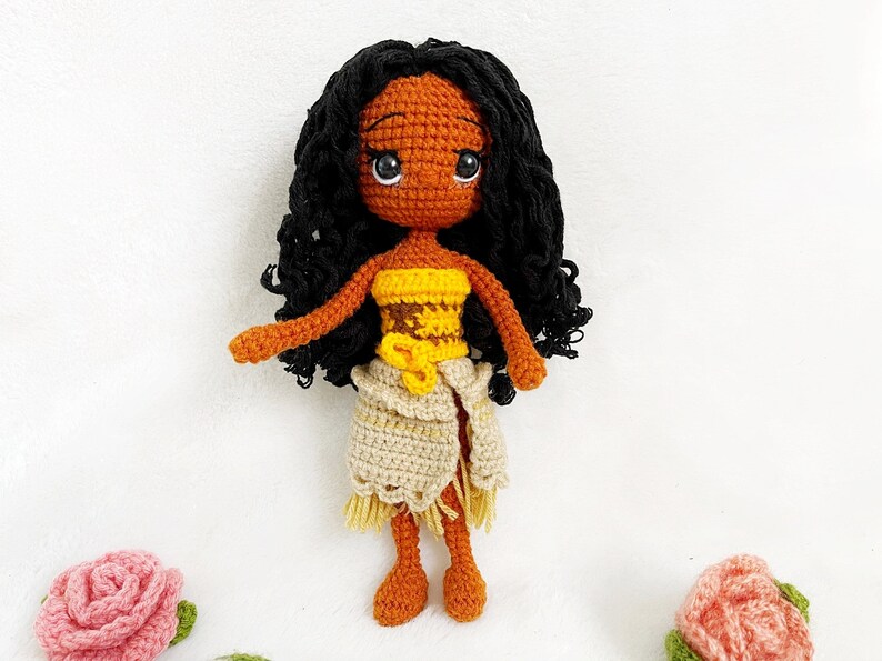 May include: Crocheted doll with brown skin, black curly hair, and a yellow and brown top. The doll is wearing a beige skirt with a fringe and is standing on a white background.
