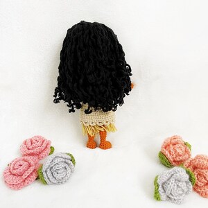 May include: A crocheted doll with long black curly hair, wearing a beige dress with a brown fringe, and orange legs. The doll is standing on a white surface with five crocheted roses in pink and gray.