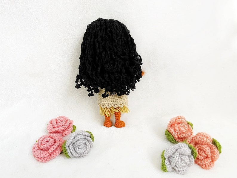 May include: A crocheted doll with long black curly hair, wearing a beige dress with a brown fringe, and orange legs. The doll is standing on a white surface with five crocheted roses in pink and gray.