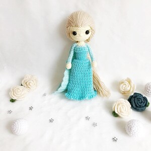 May include: A crocheted doll with long blonde hair wearing a light blue dress and a white cape. The doll is surrounded by white and green crocheted flowers and white balls.