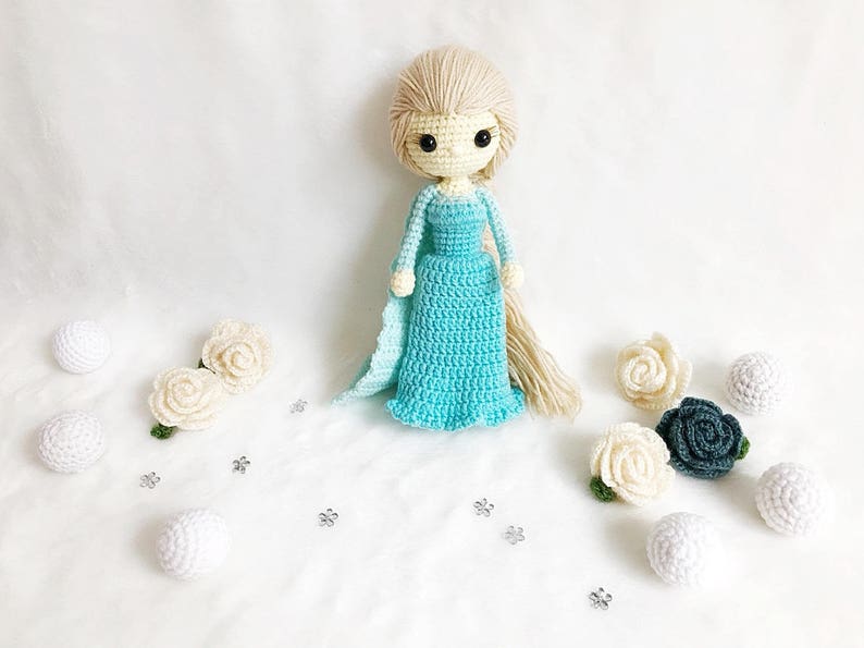 May include: A crocheted doll with long blonde hair wearing a light blue dress and a white cape. The doll is surrounded by white and green crocheted flowers and white balls.