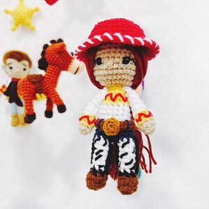 Puede incluir: Una mu&ntilde;eca de crochet vestida de vaquera. La mu&ntilde;eca tiene el pelo rojo, un sombrero rojo con ribete blanco, una camisa blanca con detalles amarillos, un cintur&oacute;n marr&oacute;n, pantalones vaqueros azules y botas marrones. La mu&ntilde;eca sostiene una cuerda roja.