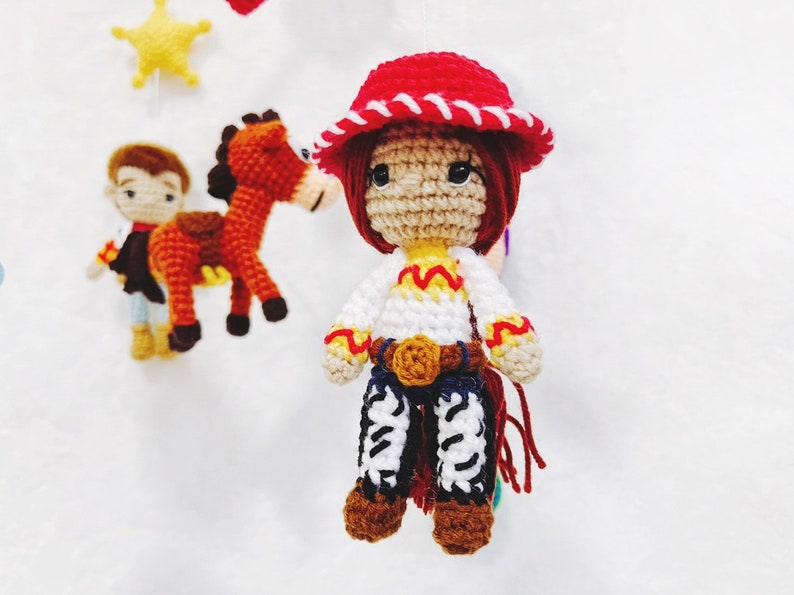 Puede incluir: Una mu&ntilde;eca de crochet vestida de vaquera. La mu&ntilde;eca tiene el pelo rojo, un sombrero rojo con ribete blanco, una camisa blanca con detalles amarillos, un cintur&oacute;n marr&oacute;n, pantalones vaqueros azules y botas marrones. La mu&ntilde;eca sostiene una cuerda roja.