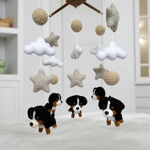 Op de afbeelding: Een handgemaakte baby mobile met gehaakte elementen. De mobile bevat beige bollen, grijze sterren, witte wolken met blauwe rand en zwarte, bruine en witte puppyfiguren. Het houten frame hangt de decoraties op.