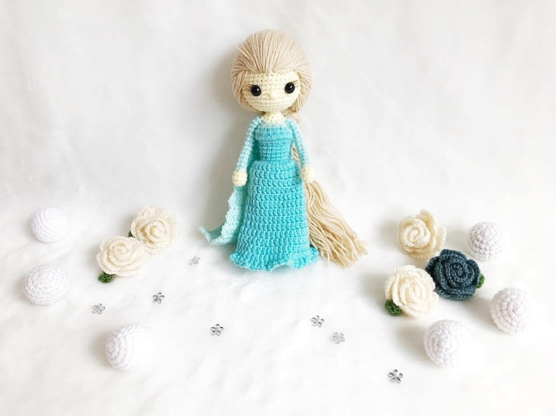 May include: A crocheted doll with long blonde hair wearing a light blue dress. The doll is surrounded by white and green crocheted flowers and white balls.