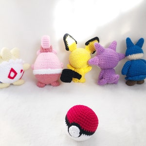 May include: A collection of six handmade crochet figures in various colors. The figures include a yellow Pikachu, a red and white Pokeball, and other characters in shades of pink, purple, blue, and white. The figures are arranged on a white surface.