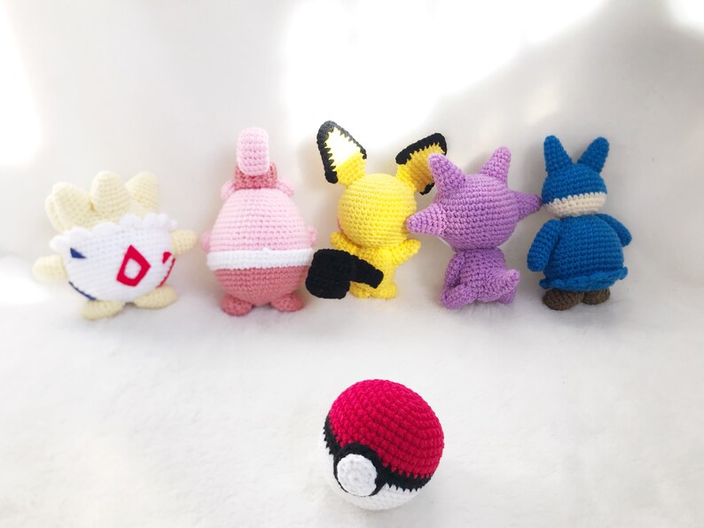 May include: A collection of six handmade crochet figures in various colors. The figures include a yellow Pikachu, a red and white Pokeball, and other characters in shades of pink, purple, blue, and white. The figures are arranged on a white surface.
