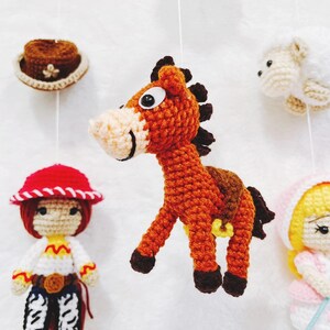 Puede incluir: Un m&oacute;vil de crochet con un caballo marr&oacute;n, una oveja blanca, una ni&ntilde;a pelirroja con un sombrero rojo y blanco, y una ni&ntilde;a rubia con un vestido rosa y un palo azul.