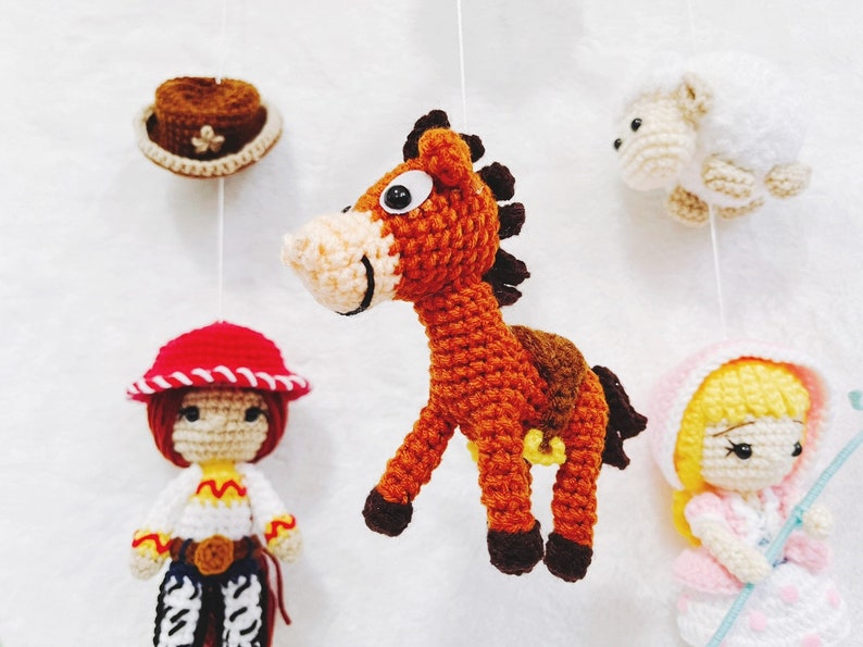Puede incluir: Un m&oacute;vil de crochet con un caballo marr&oacute;n, una oveja blanca, una ni&ntilde;a pelirroja con un sombrero rojo y blanco, y una ni&ntilde;a rubia con un vestido rosa y un palo azul.