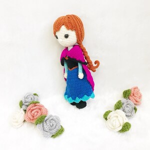 May include: A crocheted doll with brown hair, a blue dress, and a pink cape. The doll is surrounded by crocheted flowers in pink, white, and gray.