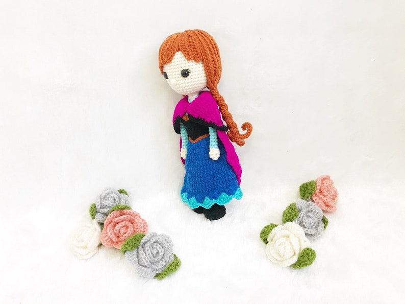 May include: A crocheted doll with brown hair, a blue dress, and a pink cape. The doll is surrounded by crocheted flowers in pink, white, and gray.