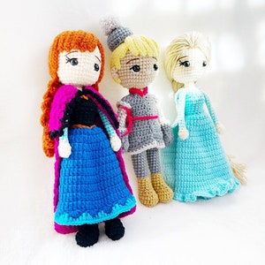 May include: Three crocheted dolls, one with red and orange hair, one with blonde hair, and one with brown hair. The dolls are wearing dresses and are standing on a white surface.