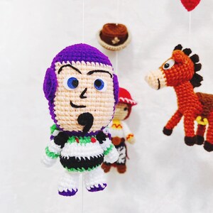 Puede incluir: Un juguete de crochet de Buzz Lightyear de la franquicia Toy Story. El juguete es morado, blanco y verde, con un casco morado, ojos blancos y una camisa a rayas verdes y blancas.