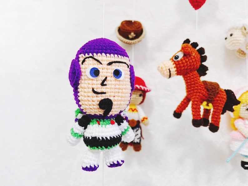 Puede incluir: Un juguete de crochet de Buzz Lightyear de la franquicia Toy Story. El juguete es morado, blanco y verde, con un casco morado, ojos blancos y una camisa a rayas verdes y blancas.