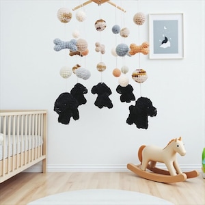 May include: A nursery mobile with crochet spheres, dog and bone shapes in neutral tones. The mobile hangs above a wooden cot and a rocking horse. The room has a neutral colour scheme.
