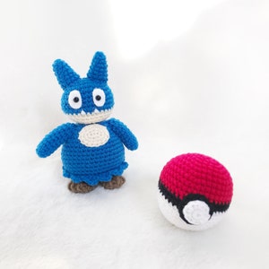 May include: A handmade blue crochet creature with a white belly and brown feet stands next to a red and white crochet ball. The creature has large eyes and pointed ears. The ball has a black stripe.