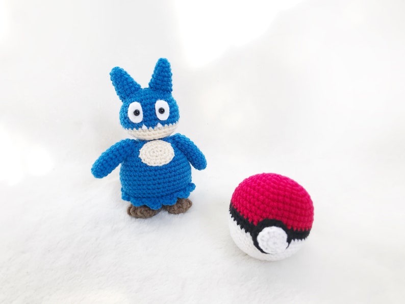 May include: A handmade blue crochet creature with a white belly and brown feet stands next to a red and white crochet ball. The creature has large eyes and pointed ears. The ball has a black stripe.