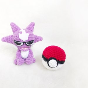 May include: A handmade crochet figure of a purple cartoon character with black sunglasses and a white emblem. Beside it is a red and white crochet ball. The items are on a white surface. The theme is related to a popular animated series.