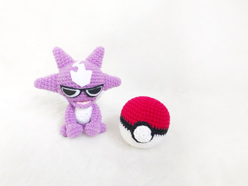 May include: A handmade crochet figure of a purple cartoon character with black sunglasses and a white emblem. Beside it is a red and white crochet ball. The items are on a white surface. The theme is related to a popular animated series.