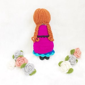 May include: A crocheted doll with long brown hair, wearing a pink dress with a blue and white trim, standing on a white surface with three crocheted flowers.