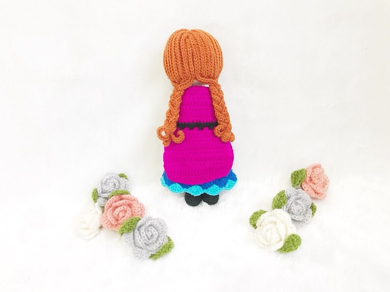 May include: A crocheted doll with long brown hair, wearing a pink dress with a blue and white trim, standing on a white surface with three crocheted flowers.