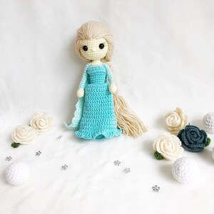 May include: A crocheted doll with long blonde hair wearing a light blue dress. The doll is surrounded by white and green crocheted flowers and white balls.