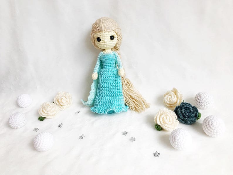 May include: A crocheted doll with long blonde hair wearing a light blue dress. The doll is surrounded by white and green crocheted flowers and white balls.