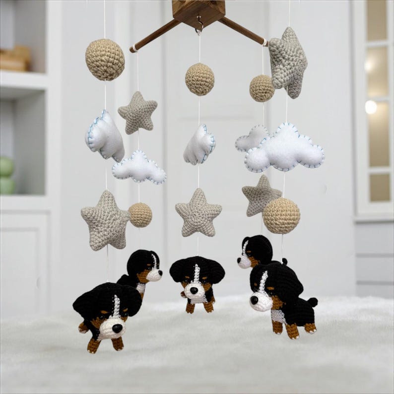 Op de afbeelding: Een handgemaakte baby mobile met gehaakte elementen. De mobile bevat beige gehaakte bollen, grijze sterren, witte wolken en schattige zwart-wit-bruine hondenfiguren. Het houten frame hangt de decoraties op.
