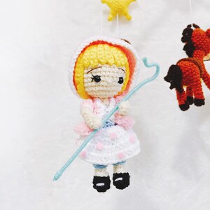 Puede incluir: Una mu&ntilde;eca de crochet con cabello rubio, vestida con un vestido rosa y blanco y sosteniendo un palo azul. La mu&ntilde;eca es parte de un m&oacute;vil con otros personajes de crochet.