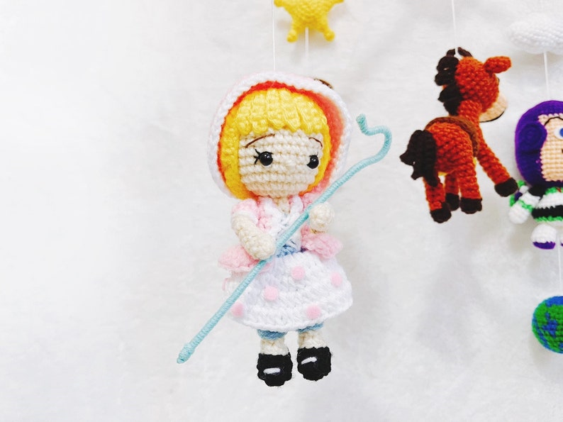 Puede incluir: Una mu&ntilde;eca de crochet con cabello rubio, vestida con un vestido rosa y blanco y sosteniendo un palo azul. La mu&ntilde;eca es parte de un m&oacute;vil con otros personajes de crochet.