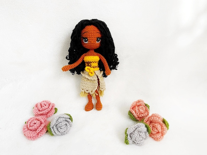 May include: Crocheted doll with brown skin, black curly hair, and a yellow and brown outfit. The doll is surrounded by crocheted flowers in pink and gray.
