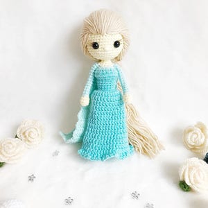 May include: A crocheted doll with long blonde hair, wearing a light blue dress. The doll has black eyes and a white face. The doll is surrounded by white crocheted flowers.