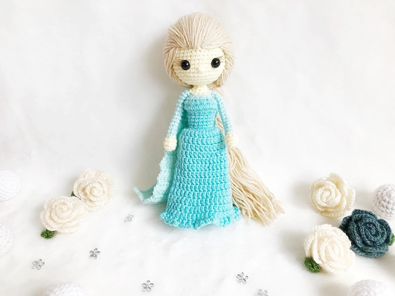 May include: A crocheted doll with long blonde hair, wearing a light blue dress. The doll has black eyes and a white face. The doll is surrounded by white crocheted flowers.